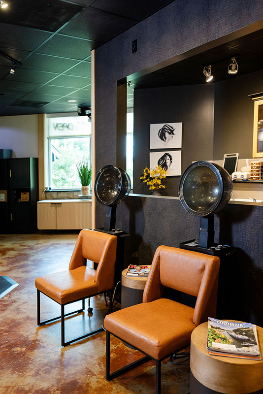 thestrand-homepage-rightchair Hair drying station at The Strand Salon and Spa in Columbia, Missouri shows faux leather chairs and a stained concrete floor with dark walls with multiple chairs.
